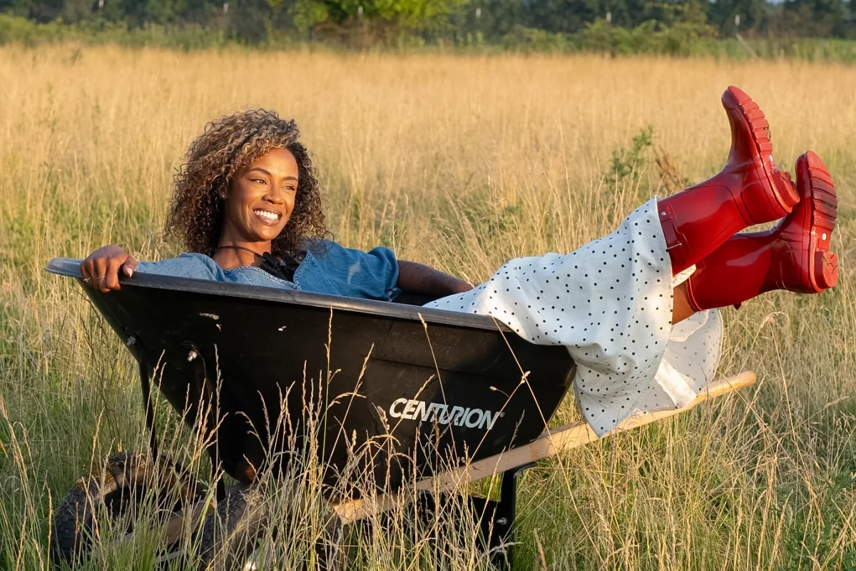 A woman smiling in a wheelbarrow with her red boots raised in a sunlit field